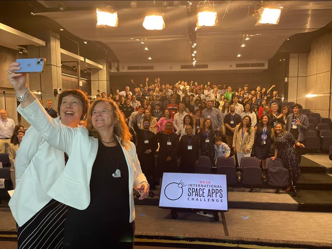 Two women smile and take a selfie onstage at an auditorium. Behind them, a large crowd stands, smiles, and waves. Credit: NASA