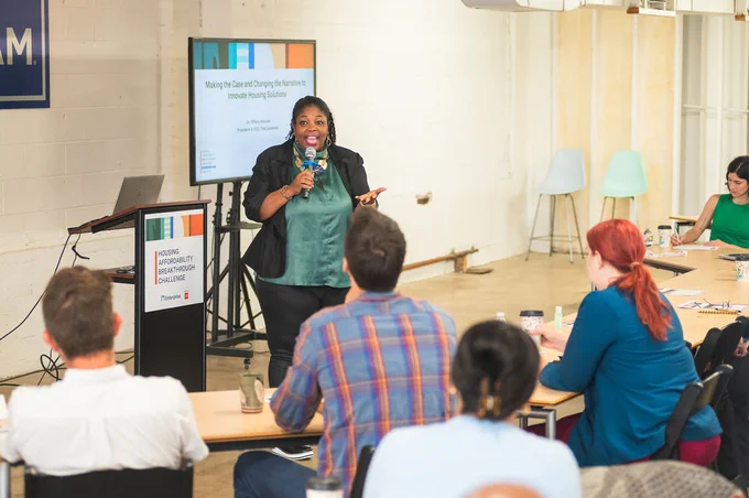 Dr Tiffany Manuel stands in front of the space where Housing Affordability Breakthrough Challenge winners are sitting, and she is holding a microphone and conducting a presentation. She’s excited and smiling widely. The room has a casual, modern setting and the audience, which is several adults seated at open and connected tables, looks engaged and interested.