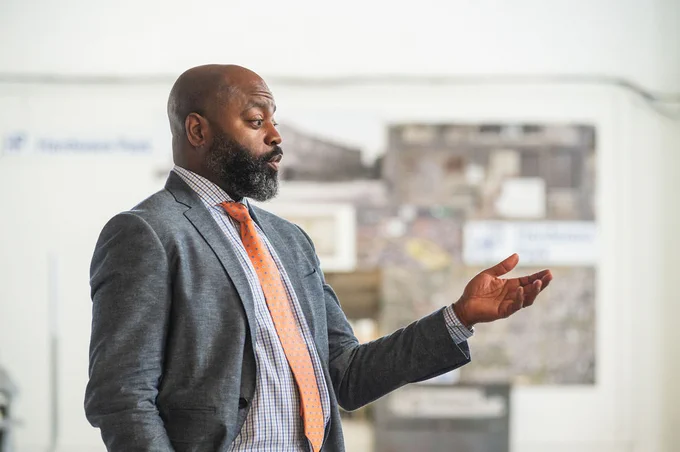 Stacy Spann, head of access and affordability at Wells Fargo stands and addresses the Housing Affordability Breakthrough Challenge winners. He looks solemn as he gestures forward with his left hand. He is wearing a gray suit and orange tie.