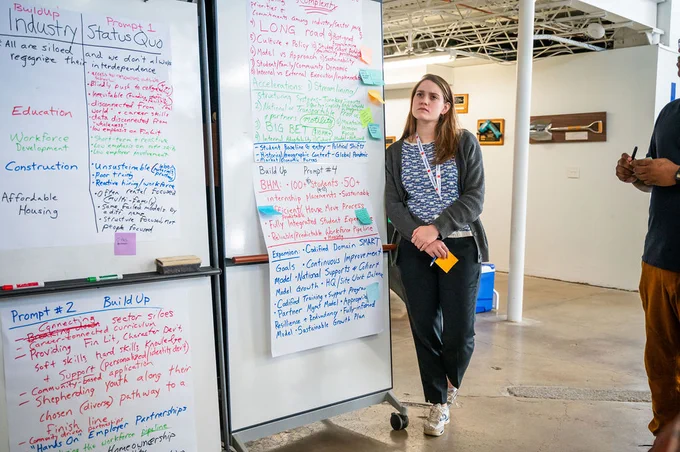 Ainsley Ash from Hope Enterprise Corporation stands and looks thoughtfully into the distance next to the group's paper posters showing their discussion and reflection and plans.
