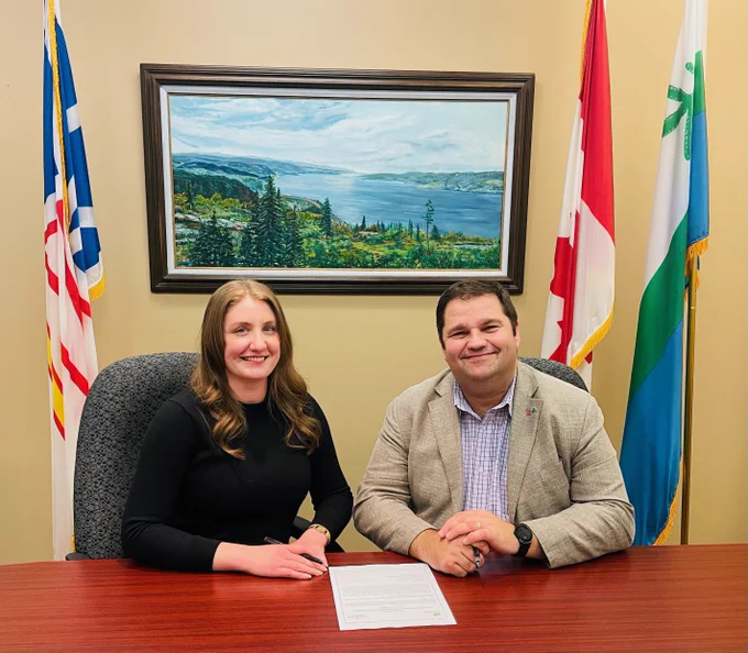 Photo of two people sitting side by side behind a desk, smiling at the camera. From left to right: Emily Cumming, Conservation Manager, Conservation Corps NL; Honourable Bernard Davis, Minister of Environment and Climate Change. 