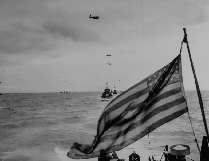 A black and white photograph of Allied ships descending upon Normandy. A U.S. flag is prominently displayed.