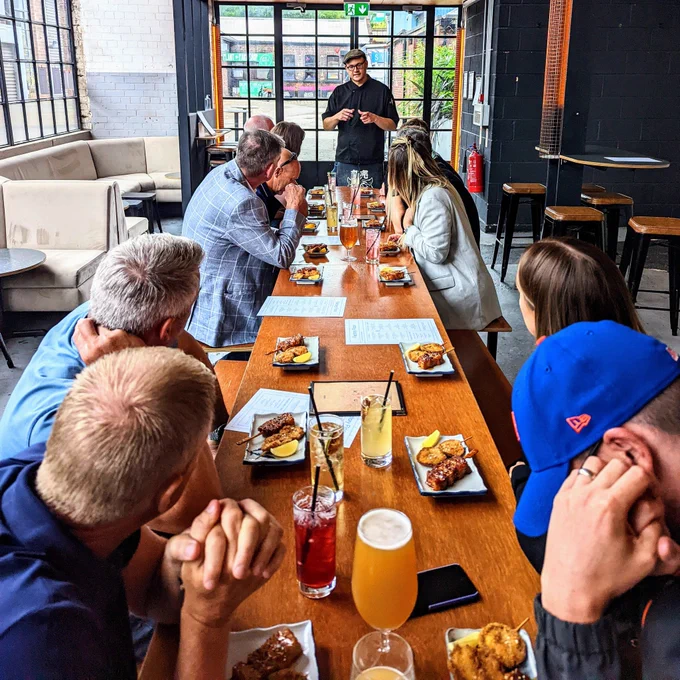 Looking down a long table set up with tasting plates, with people sat either side listening to a tour guide on a Kelham Island Food Tour