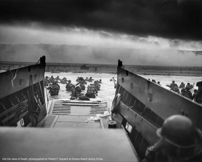 Photo 'Into the Jaws of Death' photographed by Robert F. Sargent at Omaha Beach during D-Day. You see soldiers exiting the boat towards the beach, a dark sky and smoke in the air.