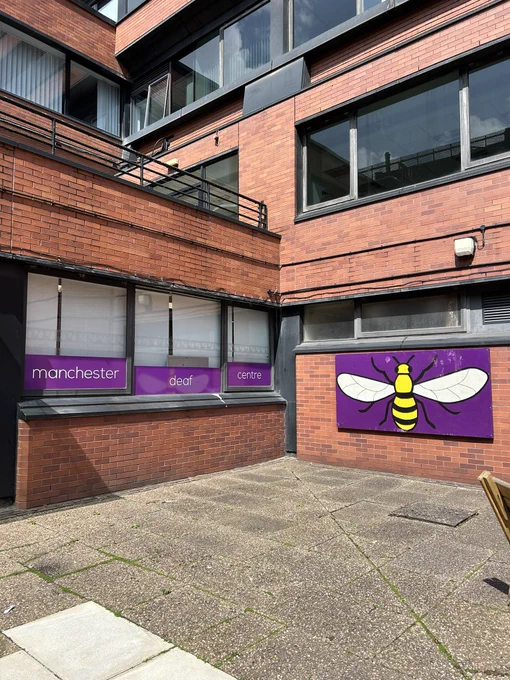 Stone paving stones leading over to a red brick building. On a series of three windows purple banners with white text read: Manchester deaf centre. On the wall attached to this is another large purple banner with a bee depicted on it.