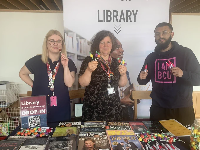 A photo of three people stood behind a table covered in books & journals, with a bennet behind them saying “Library” one person is holding library pens & the other two people lolly pops