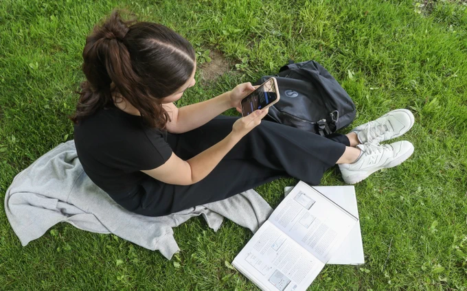 A student sitting on the grass looks at her cell phone, with backpack and books next to her