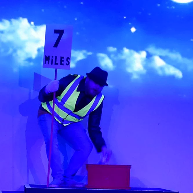 An actor playing a route marshal in Cheshire Cats. He holds a sign saying 7 miles.