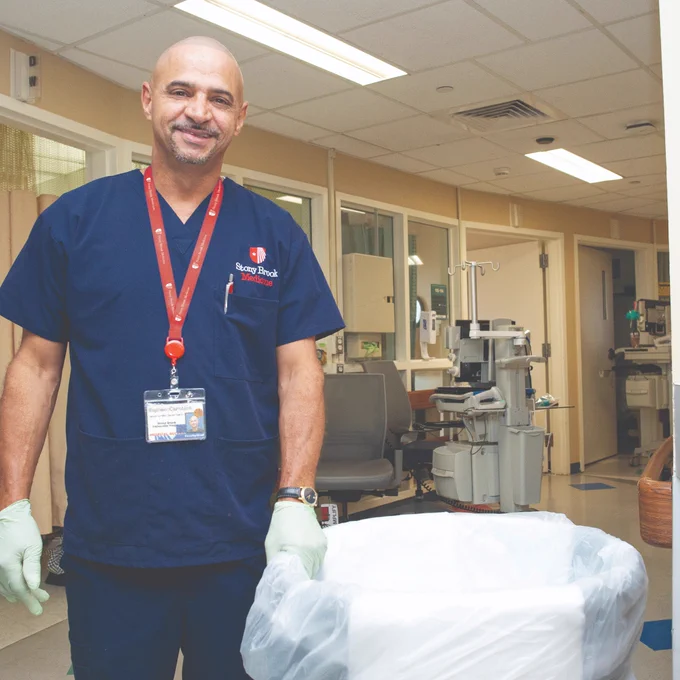 Medical professional in navy blue scrubs and gloves standing in a hospital hallway, smiling at the camera. The individual is wearing a badge that reads "Stony Brook Medicine." A hospital bed and medical equipment are visible in the background.
