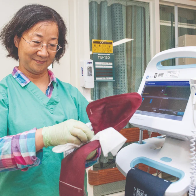 A healthcare professional in scrubs carefully cleans a medical device in a hospital setting, with various medical equipment and curtains visible in the background.