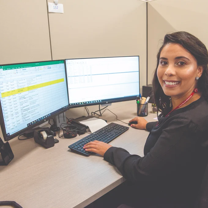 Professional at a desk in an office, smiling at the camera while working on a spreadsheet on a computer.