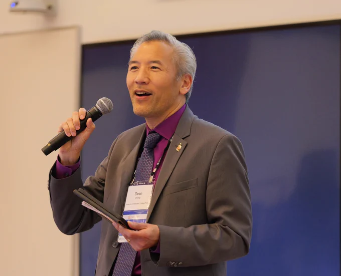 A person in a business suit holding a microphone and a tablet, speaking at a conference. There is a blue screen in the background. The speaker has a name tag labeled "Dean.“