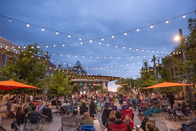 A crowd of people sit in chairs at tables at Old Town Fort Collins, Colorado as a band plays on a stage in the background. Some people are dancing near the stage. 