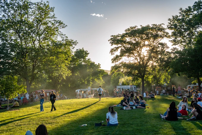 Members of the Colorado State University community gather on the grass under the shade of large, leafy trees to eat food they got at one of the food trucks lined up at the FoCo Food Truck Rally.