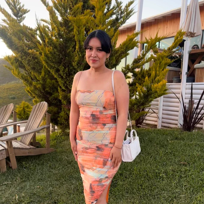 A young woman in an orange dress stands in front of a house and greenery.
