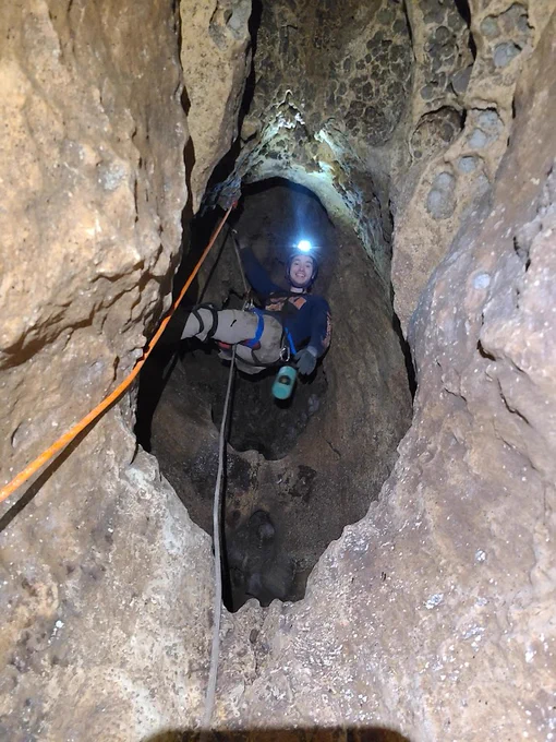 A man in caving gear hangs from a rope in a harness with a headlamp on