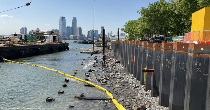 Construction site by a river with a city skyline in the background.