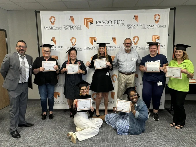 A group of eight individuals standing and kneeling in a room, all wearing graduation caps and holding certificates. They are posing in front of a backdrop with logos of "Pasco EDC," "Workforce Connect," and "SMARTstart." A man in a gray suit stands on the far left, and a woman in a green shirt stands on the far right. Two women are kneeling in the front row, while the others stand behind them, smiling for the photo.