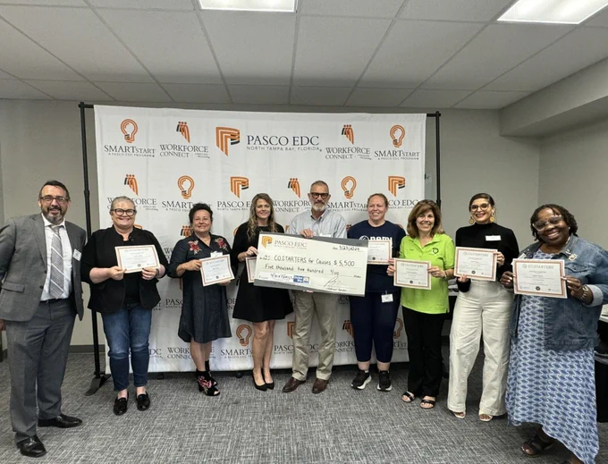 he same group of eight individuals, this time holding a large check for $5,500 along with their certificates. They are standing in front of the same backdrop with logos of "Pasco EDC," "Workforce Connect," and "SMARTstart." The man in the gray suit is on the far left, and the woman in the green shirt is holding the check with a woman in a black dress. The group is smiling and posing for the photo.