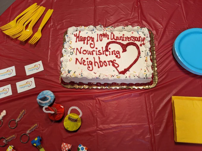 A cake on a tablecloth surrounded by party decorations. The frosting says “Happy 10th Anniversary Nourishing Neighbors”