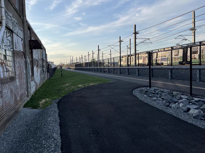 Off/on-ramp leading from/to a multi-use path. Beside the pedestrian/cycling path is a fence separating it from railroad tracks. An MBTA Green Line train passes in the background.