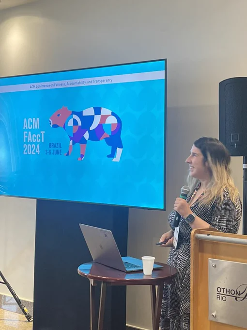 Kate stands in front of a FAccT 2024 screen that shows an artistic capibara. Kate is holding a mic.