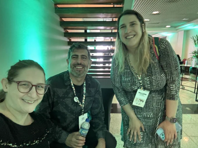 Kate, Shiri, and Foad take a photo in a large hallway underneath a tall staircase. Kate is wearing a black and white speckled dress.