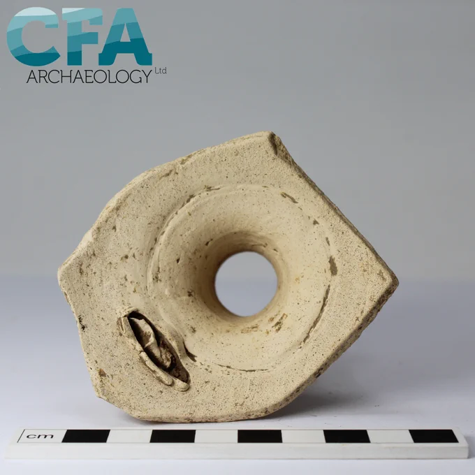 A view of the inside of a sandy white flagon, taken from underneath where it has broken just below the shoulders of the body. A slit in the body is visible, where the handle has been pushed through from the outside. It sits on a grey background with a black and white scale in front of it.