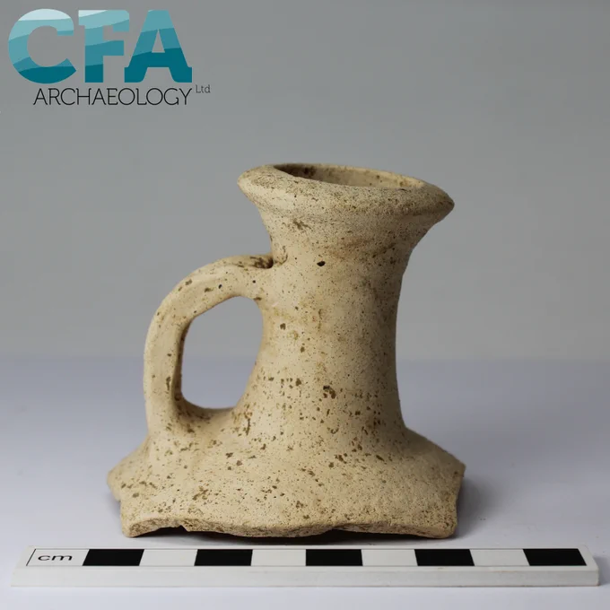 A front view of a sandy white flagon, with the neck, handle, and very top of the body visible. The rest is broken and lost. It sits on a grey background with a black and white scale in front of it. 
