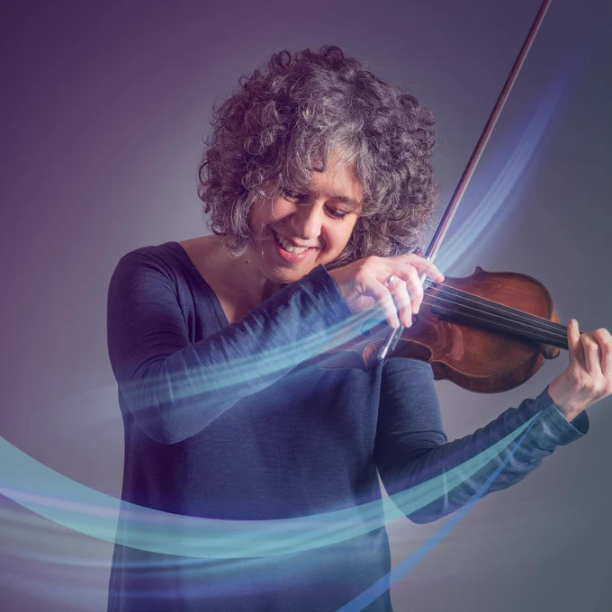 A lady smiling whilst playing the violin. She has grey curly hair and wears a lilac, long sleeve dress.