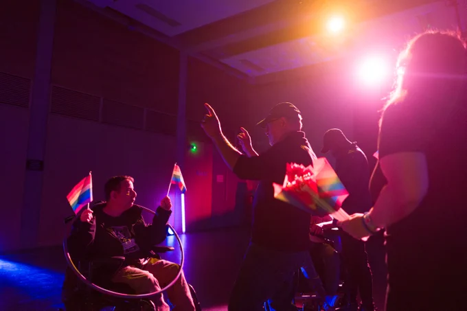 There are four people in this photo, one person is sat in his wheelchair to the left of the photo. He is waving a rainbow pride flag in each hand and has a colourful hula-hoop around his body. His support worker is stood in front of him encouraging him to wave his flags. 

There is a person stood in the background and another in the foreground with more flags.