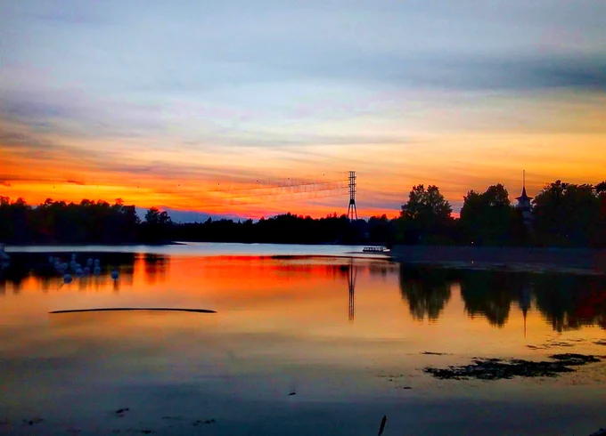 Maisema, jossa on peilityyni meri, taustalla taivasta ja puita. Aurinko on laskenut ja värjää taivaan ja meren oranssiksi.