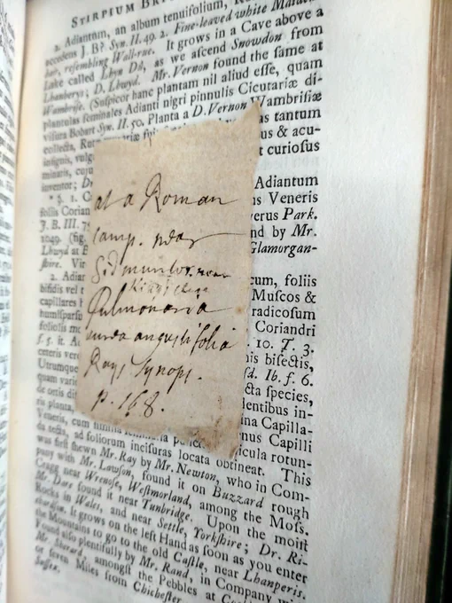 A book opened to show one page of printed Latin text and a scrap of paper bearing a handwritten note.