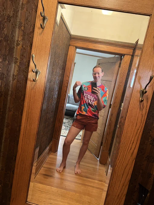 Joel in orange running shorts and a multicolored Sioux Falls City FC Pride jersey while taking a selfie in the mirror.