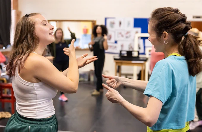 Two women, one in a white tank top and the other in a blue shirt, are engaged in conversation through the use of BSL. Other people can be seen in the background rehearsing.