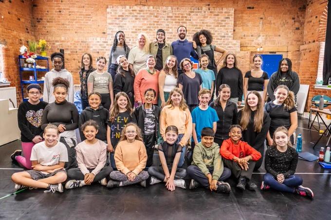 A diverse group of young people and adults pose together for a group photo in a rehearsal space with brick walls. They are smiling and look enthusiastic.