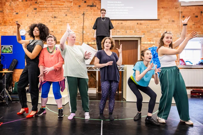 A group of actors perform a scene, each striking dynamic poses. They are in a rehearsal space with brick walls and various props and instruments around them.