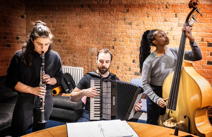 Three actor musicians are playing their instruments: a woman on the clarinet, a man on the accordion, and another woman on the double bass, all in a cozy brick-walled rehearsal room.