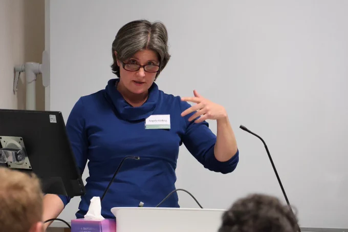 Speaker Angela Kölling delivers their paper at the conference. Looking directly at the camera over the rim of their glasses, Angela is captured mid-gesture and stands in front of a PC and microphone.