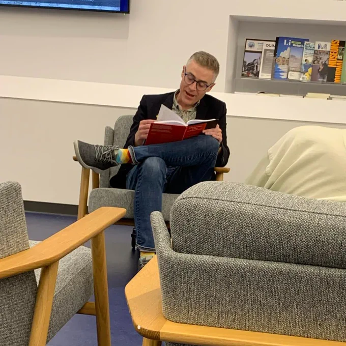 Ryan MacNeil sitting comfortably in a chair, in jeans, a patterned shirt, navy suit jacket, rainbow check socks, one leg crossed, reading from his book.