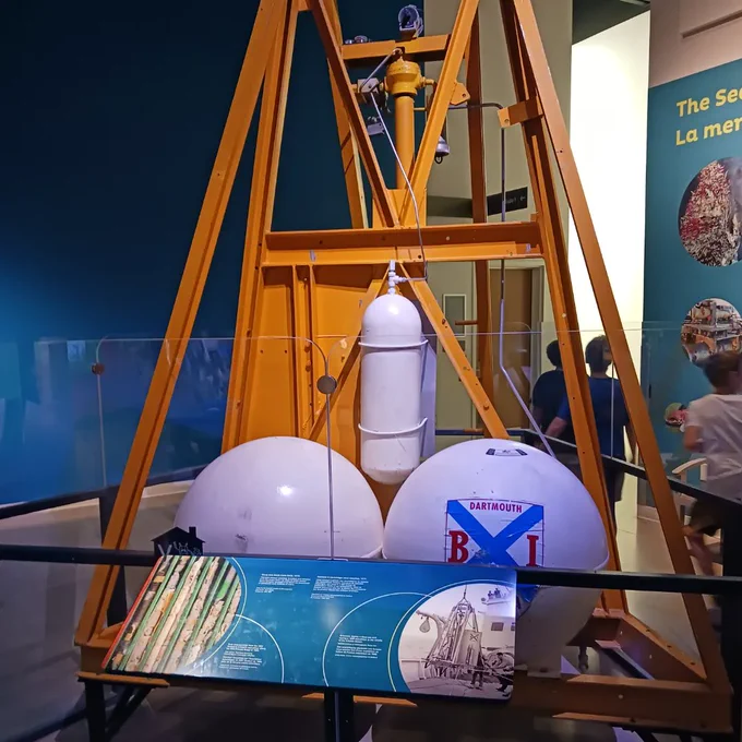 A large yellow metal tripod of at least 10 feet tall, with 2-3 diameter white metal buoyancy balls, and air cylinder, and central drill system. One of the white balls is labelled B/I Dartmouth Nova Scotia with a blue Scottish cross between the letters/words. The object is clearly an artifact in a museum and is surround by a short plexiglass wall to prevent climbing.