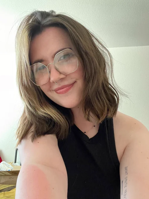 a selfie of me in even indoor lighting. my hair is slightly messy rather than neatly parted and is slightly wavy. my makeup is done but is mostly obscured by my hexagon glasses. i am wearing a black ribbed tank top and the tattoo on my arm that says “do every stupid thing” over an illustration of a match stick is visible 