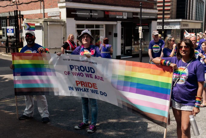 Three individuals walking in a Pride parade, holding a large rainbow flag with the inscription "PROUD OF WHO WE ARE, PRIDE IN WHAT WE DO." They are wearing colourful outfits and accessories.