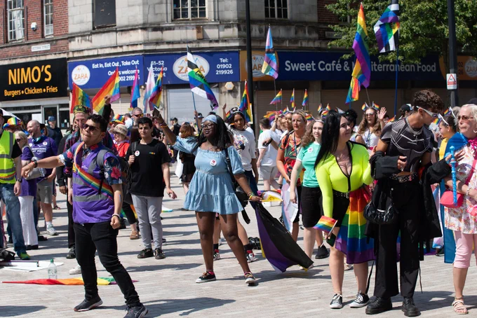 Groups of people participating in Wolverhampton Pride 2023 on a sunny day, with various rainbow flags prominently displayed. Some individuals are wearing colourful attire. Signs and banners with messages of LGBTQ  support can be seen around the area.