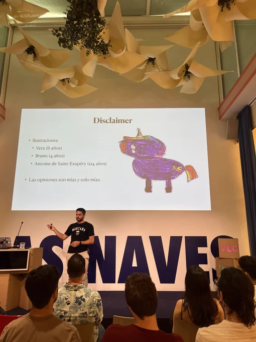 Jordi in front of his slide with the following content “Disclaimer
• Ilustraciones:
• Vera (6 años)
• Bruno (4 años)
• Antoine de Saint-Exupéry (124 años)
• Las opiniones son mías y solo mías.” and unicorn drawing