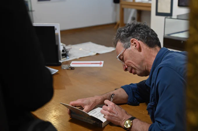Richard Askwith, a middle-aged white man wearing glasses and a blue jumper, signs a copy of his book Feet in the Clouds in the Running Int Fells exhibition in The Armitt museum. People gather in the foreground, waiting to get their books signed