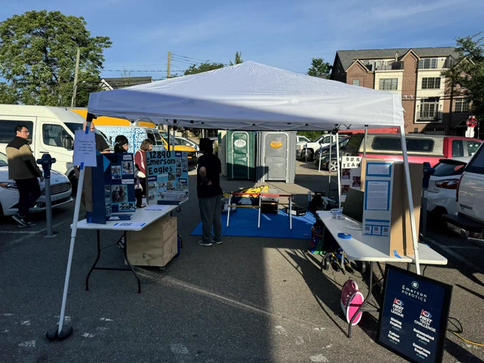 Emerson school robotics at the Ann Arbor farmers market. White canopy tent foreground. Two white folding tables with trifold poster displays. In background, blue foam flooring (on asphalt) with middle school robot and competition materials.