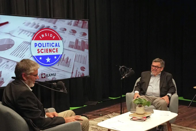 Two individuals seated on a stage engaged in conversation, with a backdrop featuring a large screen displaying the logo of "Inside Political Science" and the KAMU logo.