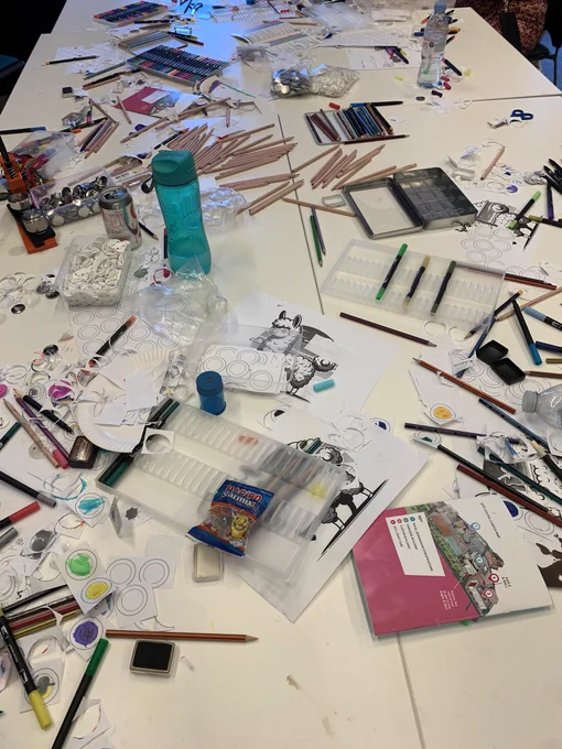 A photo of a large, white table covered in paper, colouring pens & pencils, and ink stamps - the table looks messy