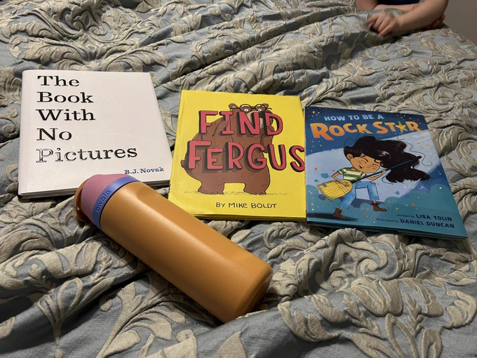 Covers of The Book With No Pictures (just the title on white background) Find Fergus (a brown bear wearing glasses hiding behind the title words)and How to be a Rockstar (a kid with long black hair shredding on a broom “guitar”) on bedcovers with the orange water bottle my toddler brought me. Cute little hand in upper right corner.